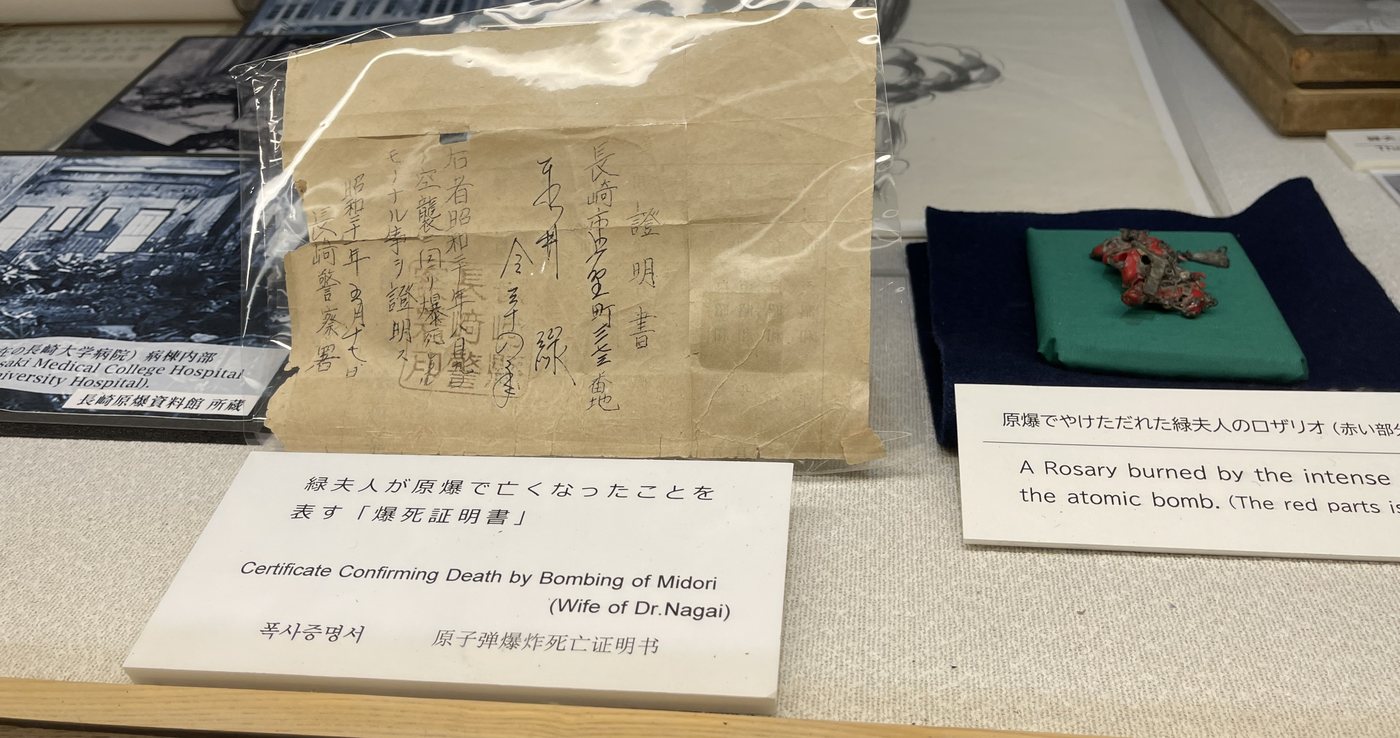 Museum display case: on the left a folded, handwritten death certificate in a clear sleeve with its label, on the right the burned rosary on a green cushion with its label